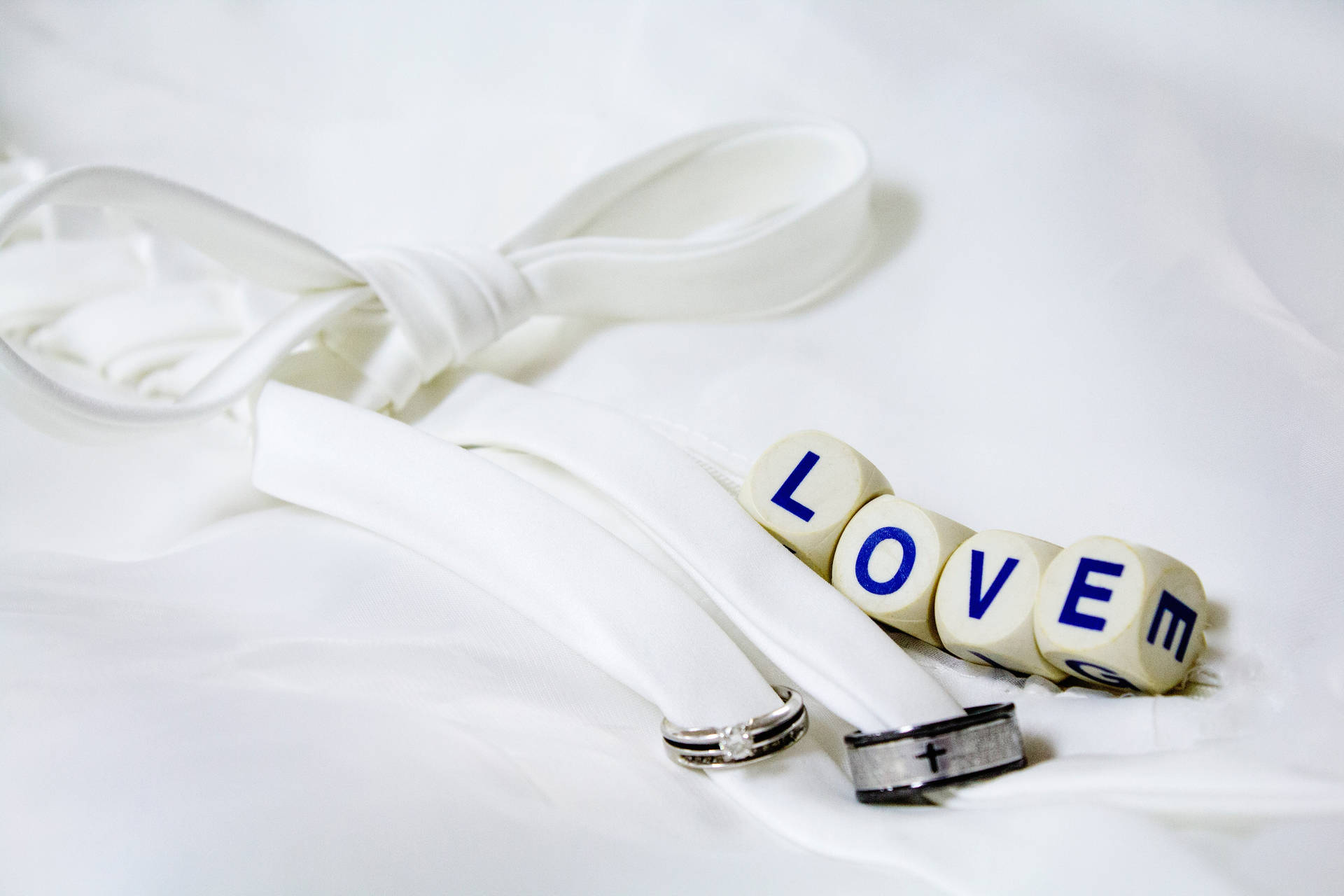 흰색 시트 위에 흰색 리본으로 묶인 은색과 검은색 웨딩 반지 옆에 'Love'라고 적힌 네 개의 흰색 주사위